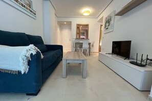 A modern living room with a blue sofa, a wooden coffee table, a TV on a white stand, and a dining area in the background.