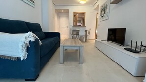 A modern living room with a blue sofa, a wooden coffee table, a TV on a white stand, and a dining area in the background.