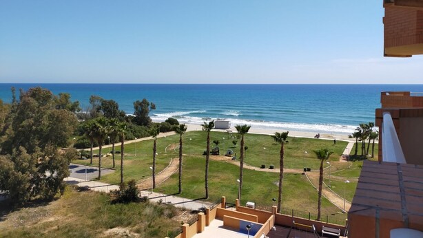 A coastal view from a balcony overlooking a beach with palm trees, a clear sky, and a calm sea.