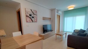 A modern living room with a wooden dining table, a flat-screen TV, a sofa, and a large window with curtains.