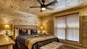 A wooden cabin bedroom with a bed, two bedside lamps, a ceiling fan, and a large window with blinds. - Hilltop Hideaway - 3 Bedroom 2 Bath - Private! (Sevierville)