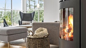A modern living room with a large window, a sofa, a chair, a woven basket, and a wood-burning stove.