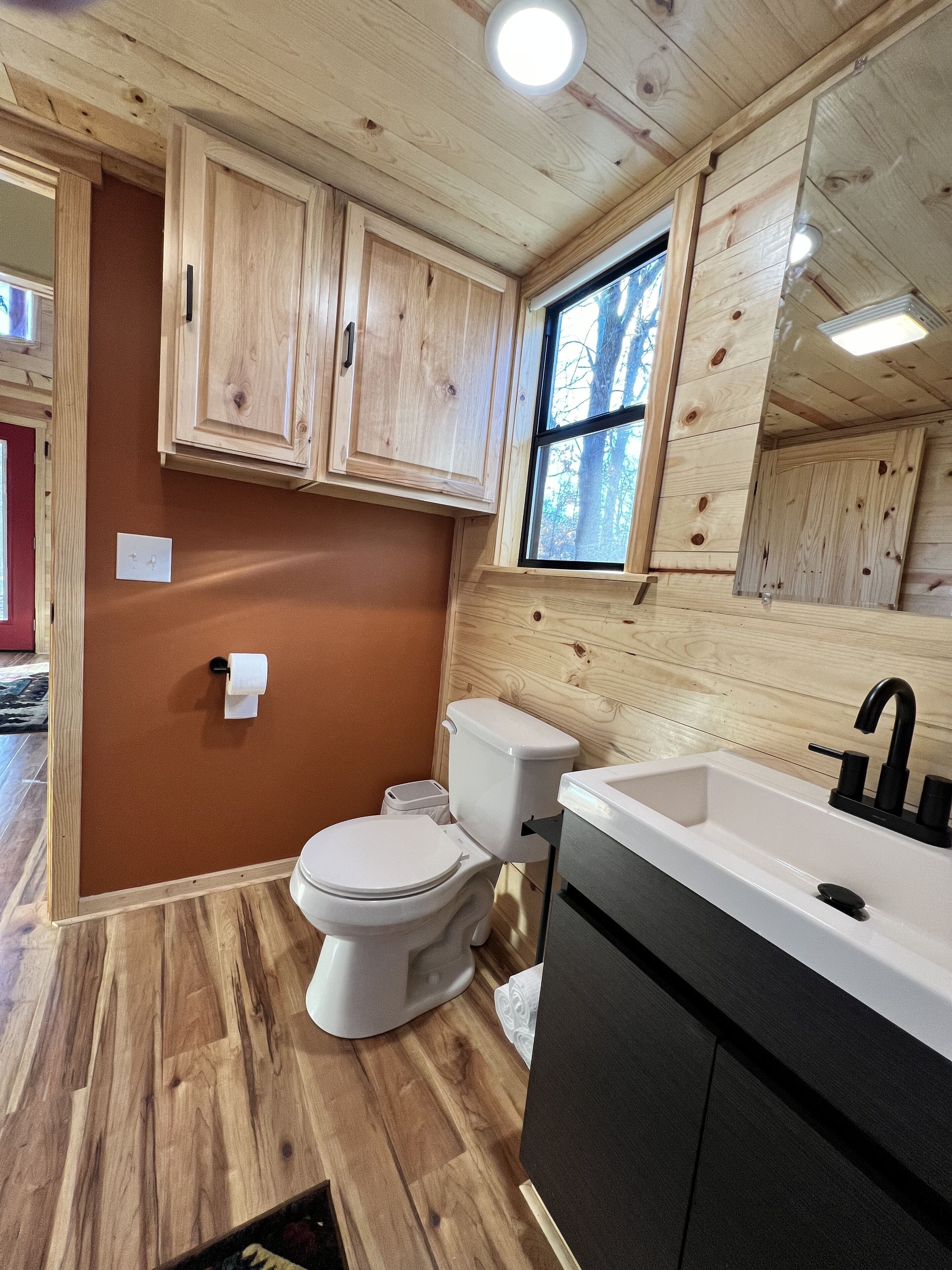 Een compacte badkamer met houten kastjes, een wit toilet, een wastafel met zwarte kraan en een raam dat voor natuurlijk licht zorgt.