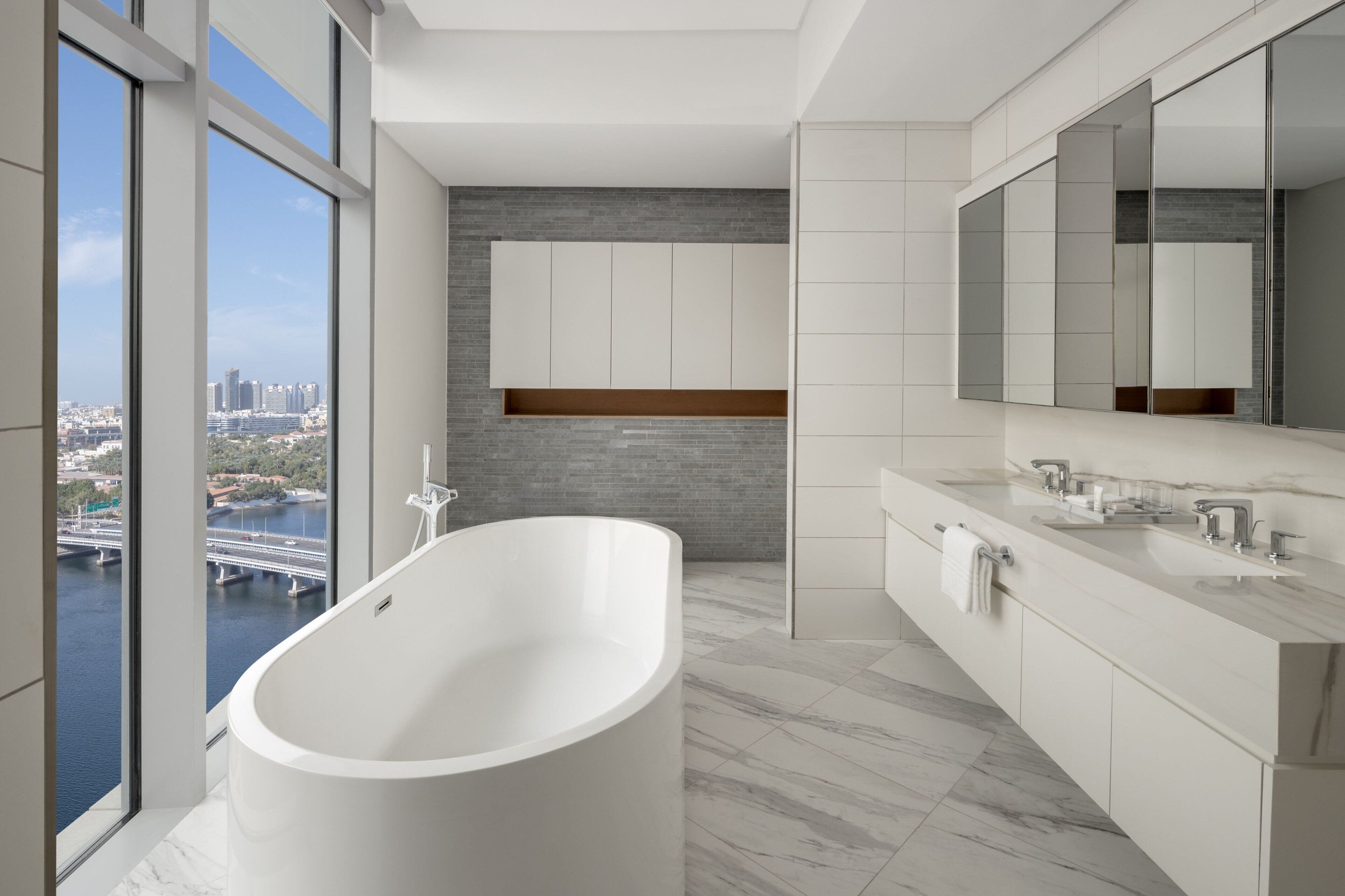A modern bathroom with a freestanding bathtub, a large mirror, and a city view through large windows.
