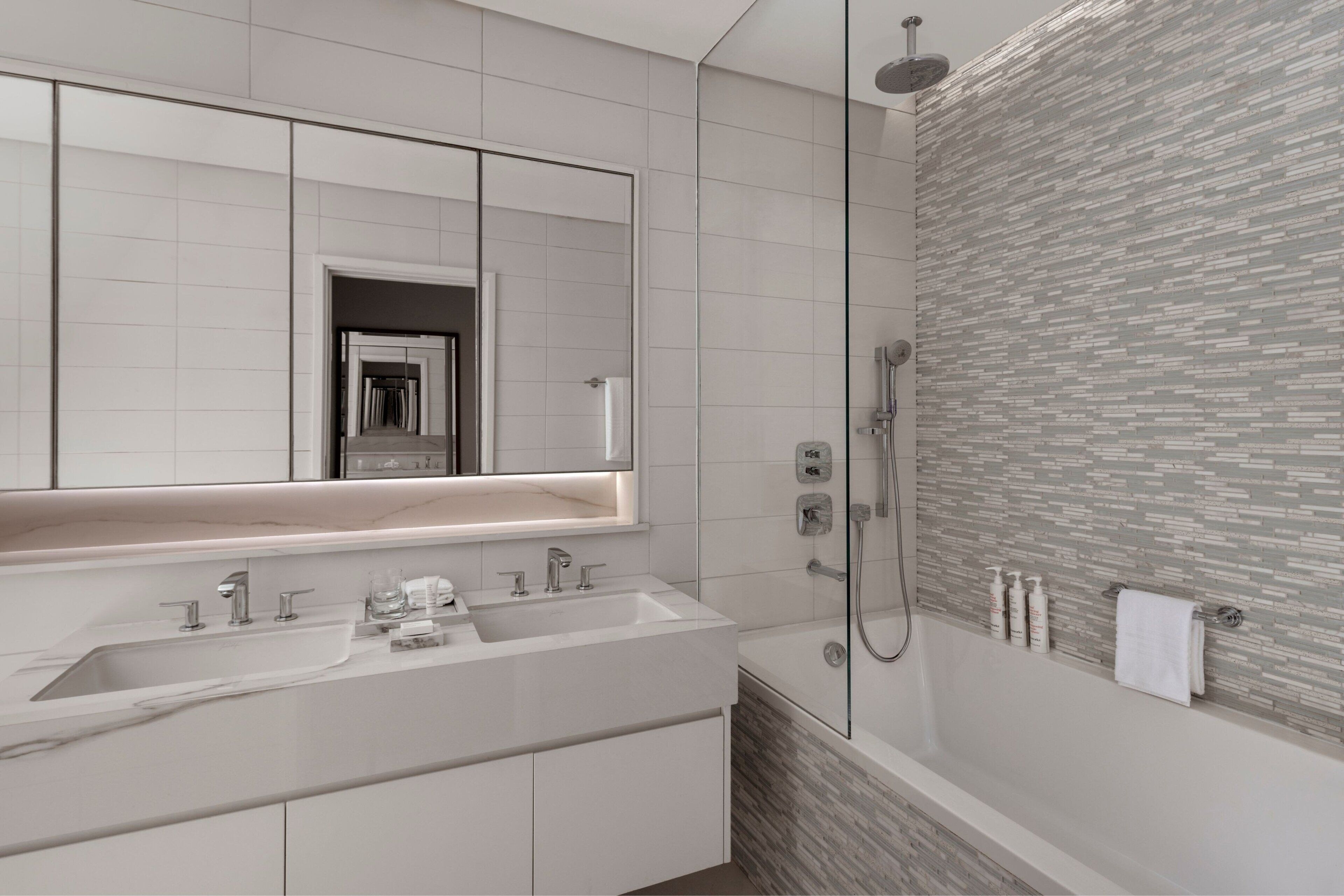 Een moderne badkamer met een grote spiegel, twee wastafels, een douche en een betegelde wand.