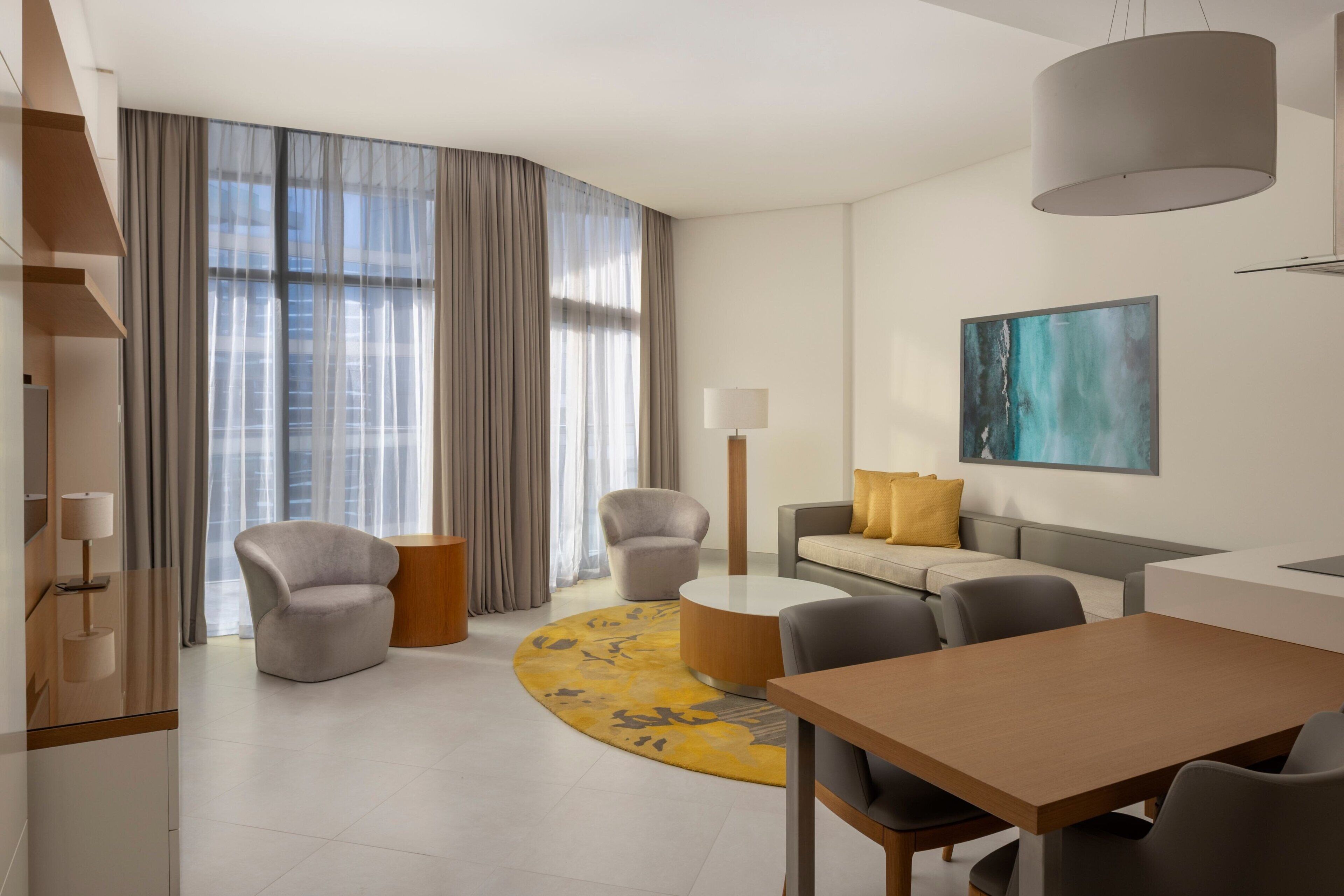 Une chambre d’hôtel moderne avec un espace de vie aux lignes courbes, une table basse ronde, un canapé agrémenté d’un coussin jaune, une table à manger et une grande fenêtre avec des rideaux.