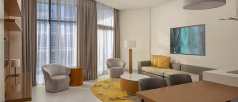 Une chambre d’hôtel moderne avec un espace de vie aux lignes courbes, une table basse ronde, un canapé agrémenté d’un coussin jaune, une table à manger et une grande fenêtre avec des rideaux.