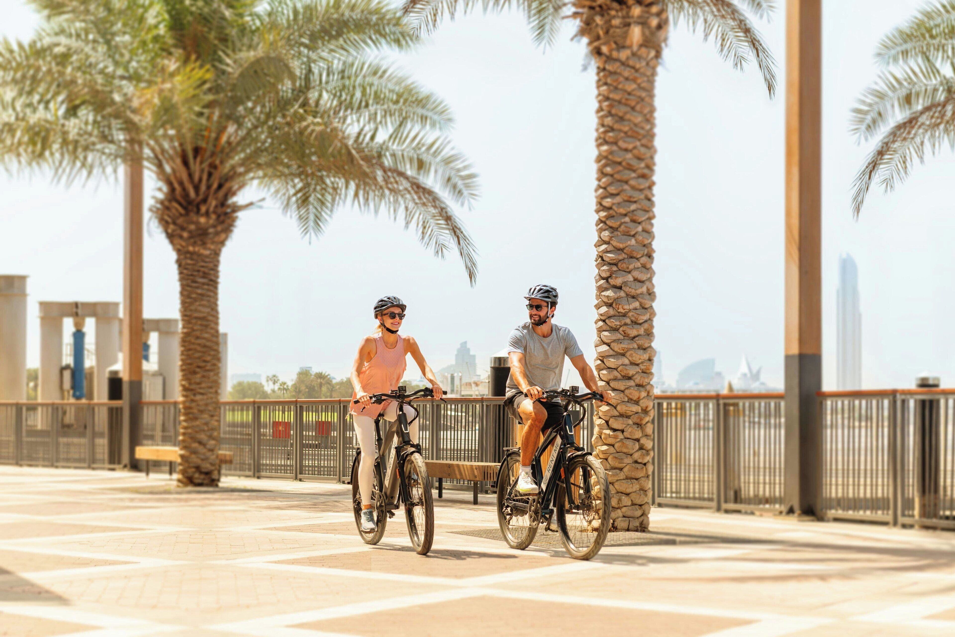 Deux personnes font du vélo sur une piste cyclable asphaltée, bordée de palmiers et d’un mur de pierres.