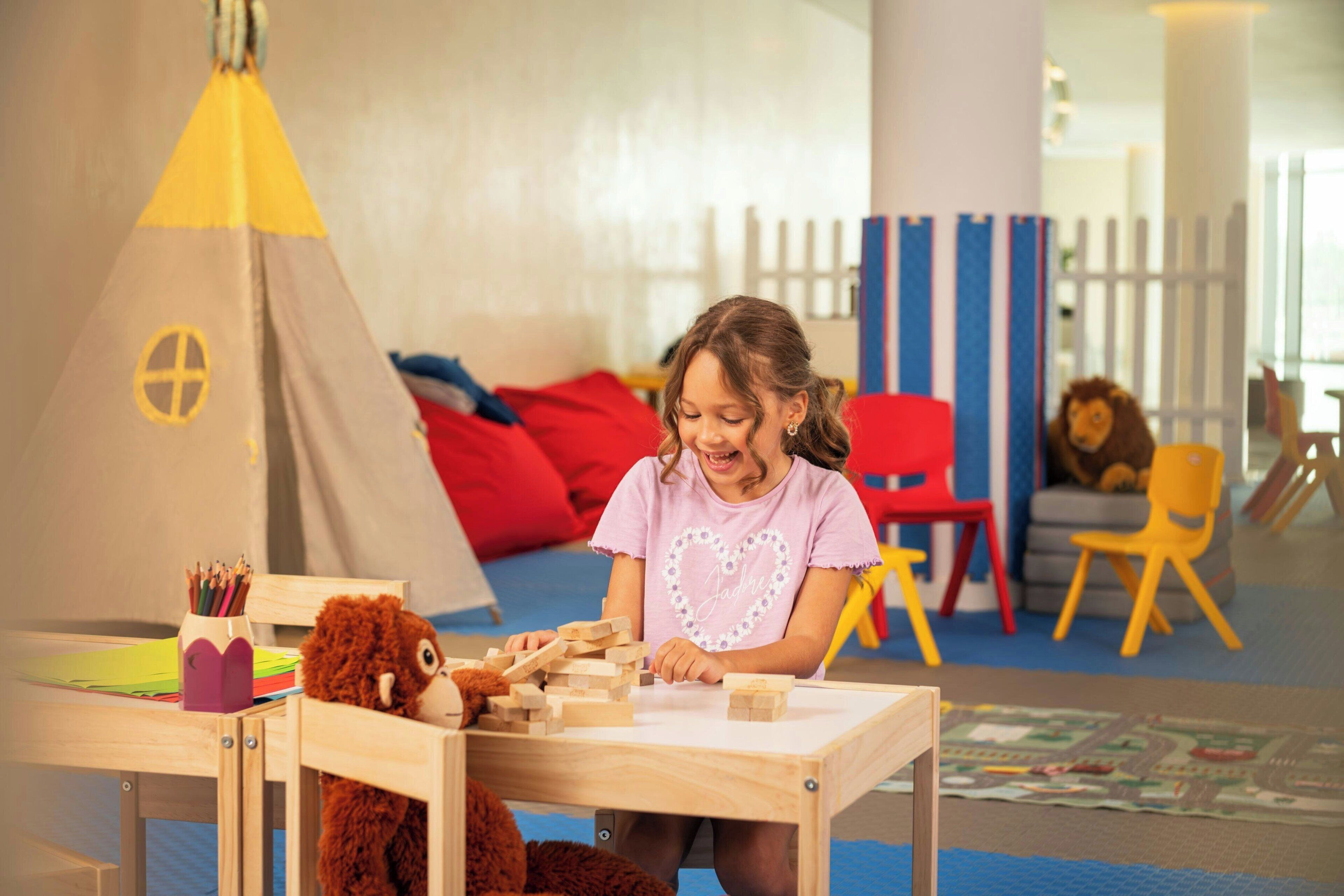 A child playing with wooden blocks at a table in a room with a play area, including a tent and colorful chairs.