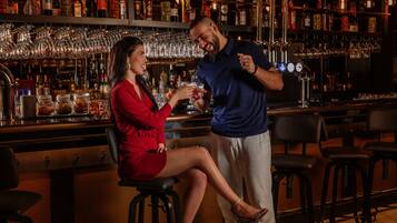 Un'area bar con un barista e un cliente. Il bancone bar presenta scaffali con diverse bottiglie e bicchieri. Sono presenti sgabelli e un bancone.
