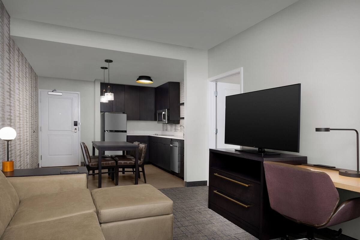 A modern living room with a sofa, a dining table with chairs, a kitchen area with dark cabinets, a flat-screen TV, and a desk with a chair.