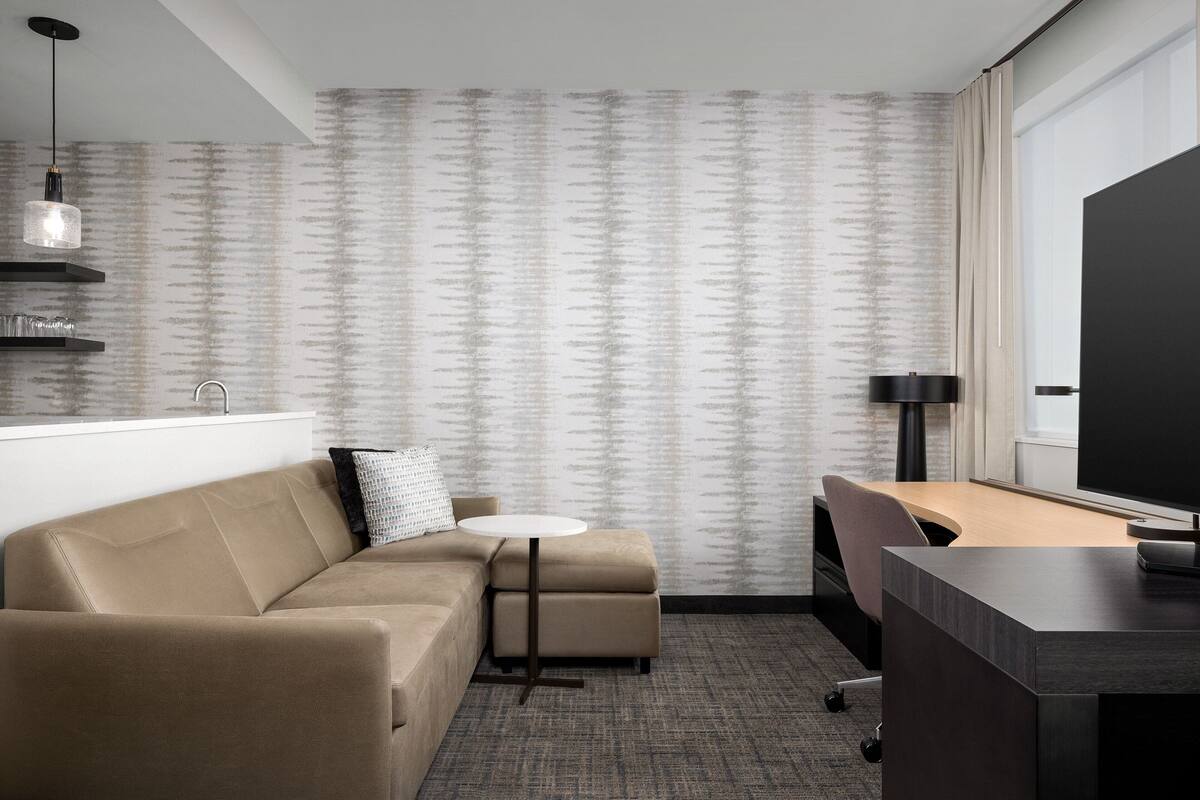 A modern hotel room with a sofa, a small round table, a desk, and a large window.