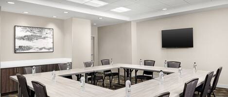 A conference room with a long table, chairs, water bottles, a flat-screen TV, and a patterned carpet.