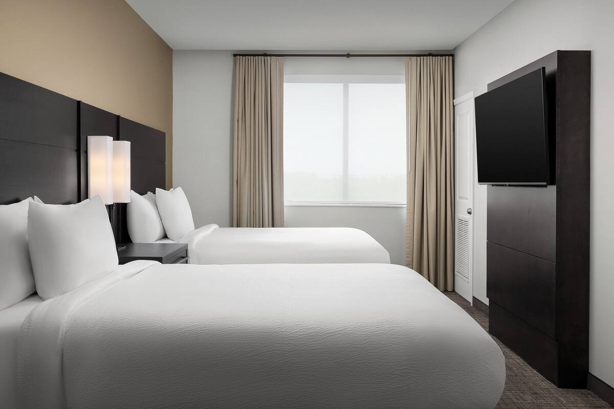A hotel room with two beds, a flat-screen TV, a nightstand, and a window with curtains.