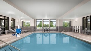 Una piscina interior moderna con paredes de vidrio transparente, agua azul y una zona de estar con mesas y sillas.