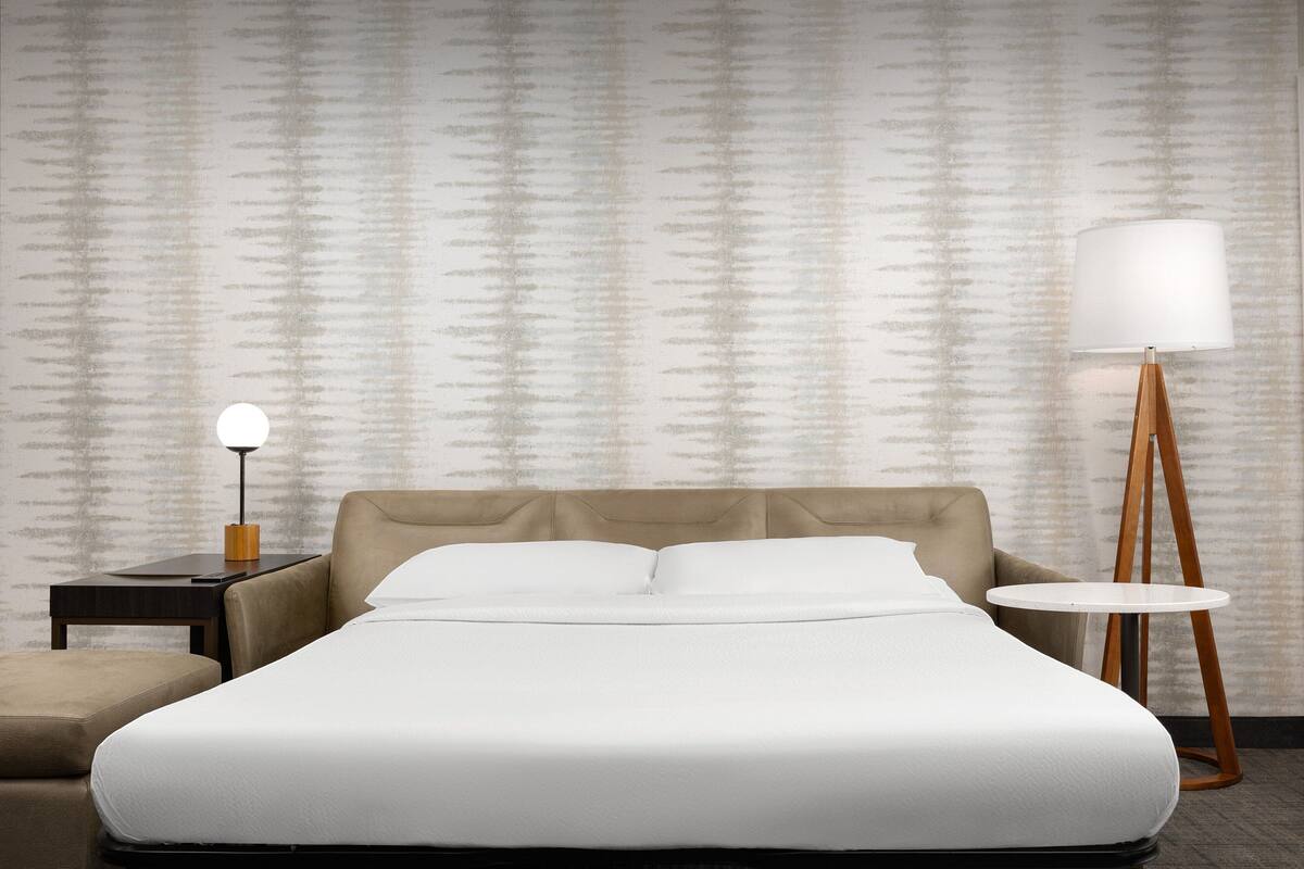 A neatly made bed with a headboard, a bedside table with a lamp, a round table, and a patterned wall.