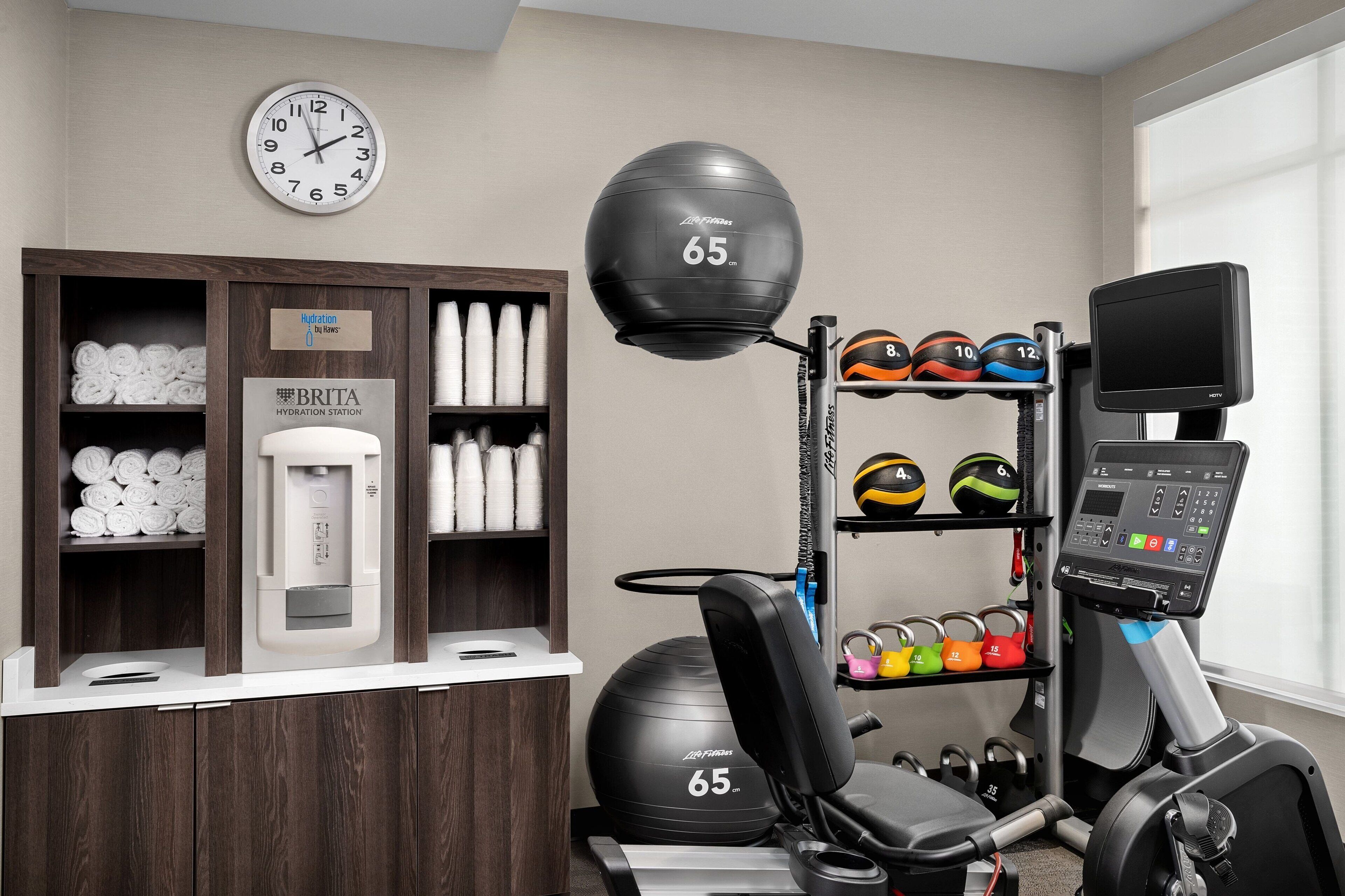 Une salle de sport équipée de matériel d’exercice, avec un point d’hydratation, ainsi que des serviettes et des bouteilles d’eau rangées avec soin.