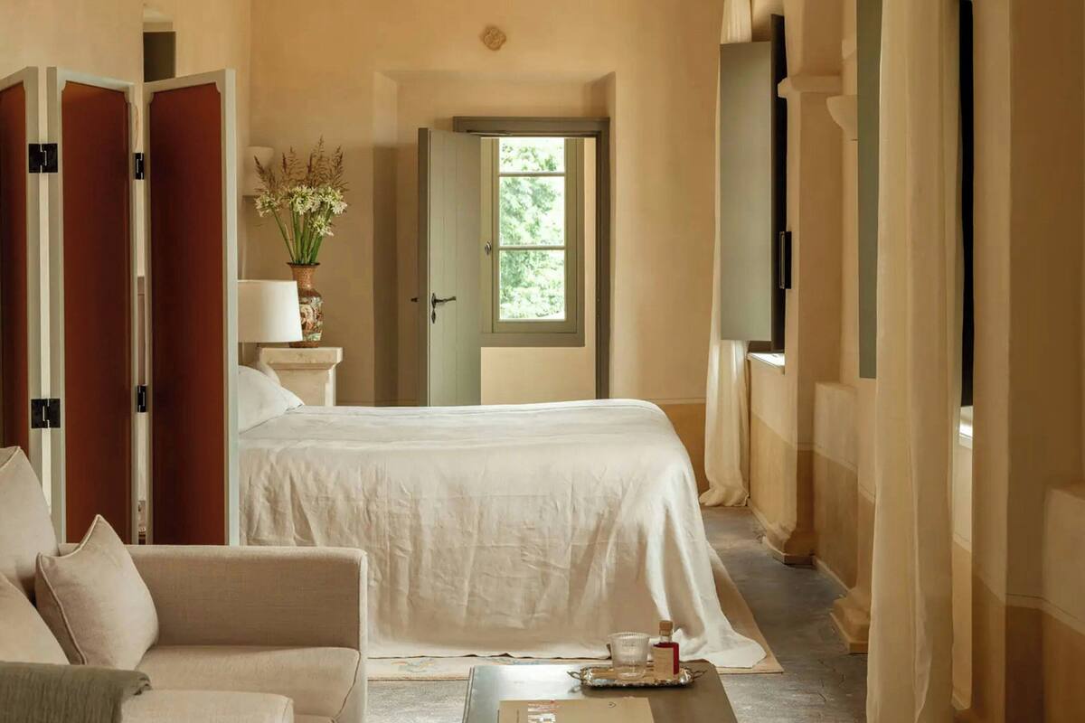 A hotel room with a bed, a sofa, a nightstand, a lamp, a vase with flowers, and a window.