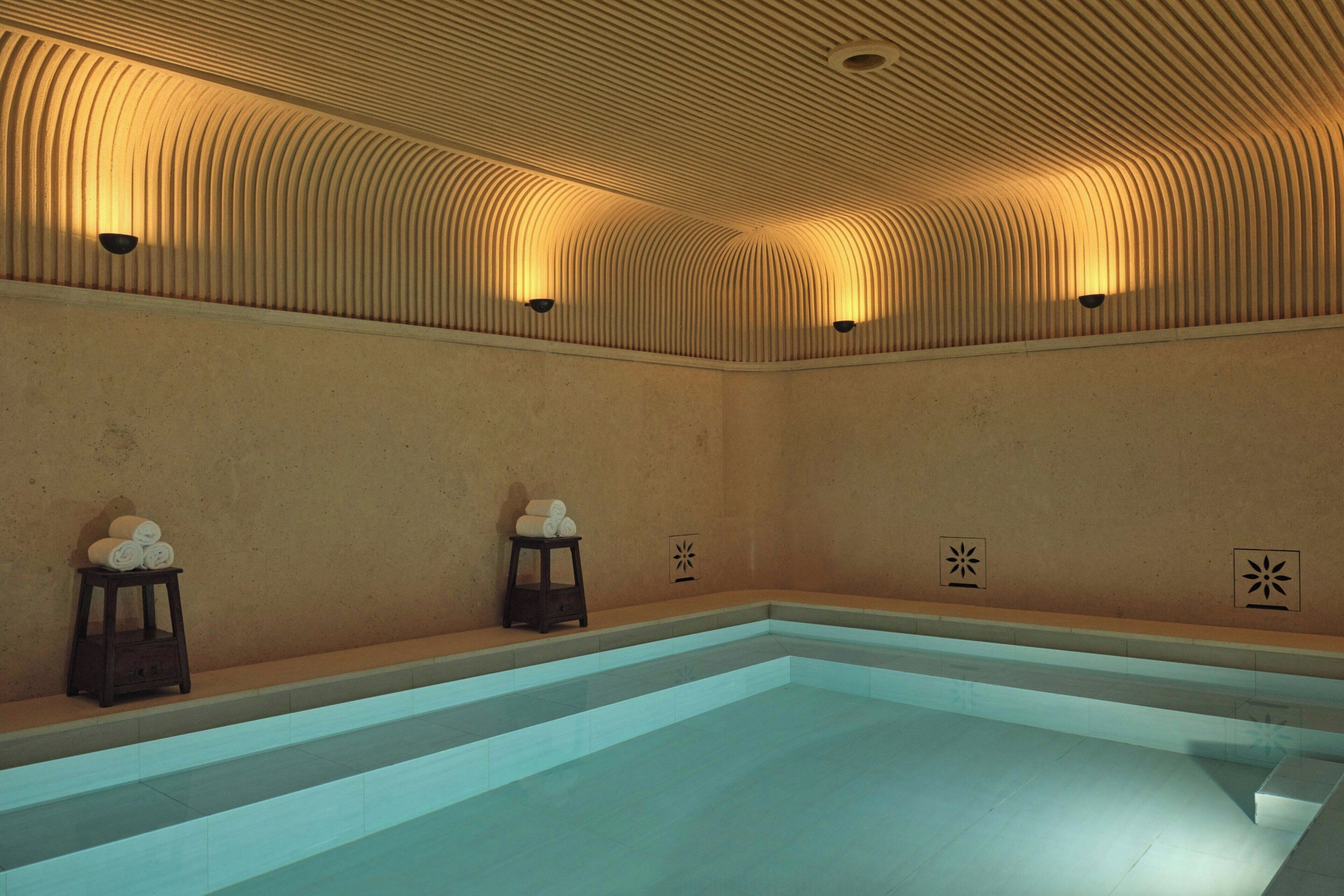 Une piscine dont le sol et les murs sont recouverts de carreaux, un plafond incurvé doté d’un éclairage encastré, et deux petits tabourets sur lesquels sont posées des serviettes.