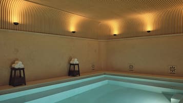 A swimming pool with a tiled floor and walls, a curved ceiling with recessed lighting, and two small stools with towels on them.