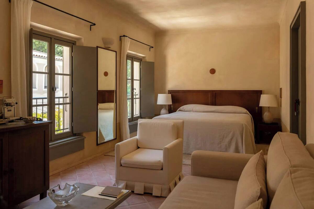 A hotel room with a bed, two bedside tables with lamps, a chair, a desk with a bowl, and a window with curtains.