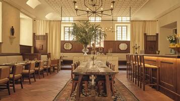 A dining area with a long wooden table, multiple chairs, a chandelier, and a large plant in the center.