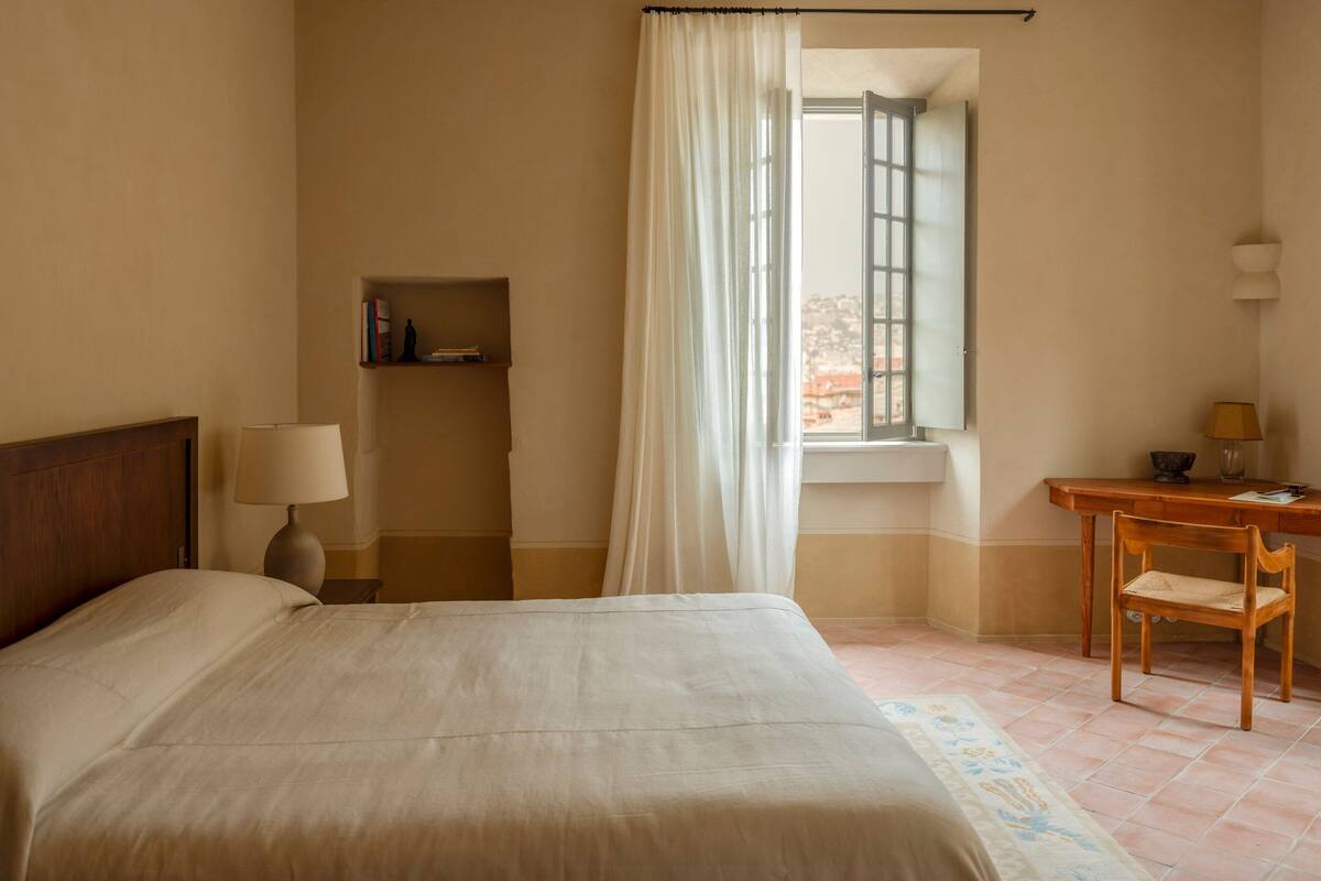 A hotel room with a bed, a desk, a chair, a window with curtains, and a small shelf with books.
