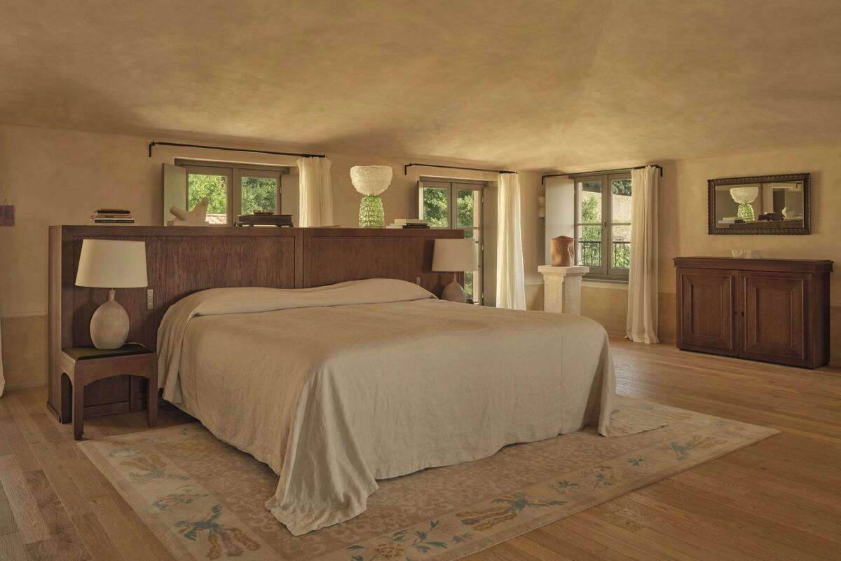 A spacious bedroom with a large bed, two bedside tables with lamps, a wooden dresser, and a view of greenery through the windows.