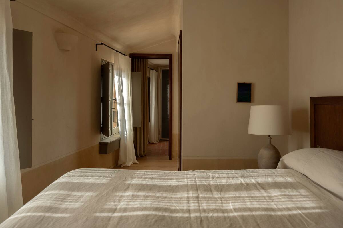 A hotel room with a bed, a nightstand with a lamp, a window with curtains, and a door leading to another room.