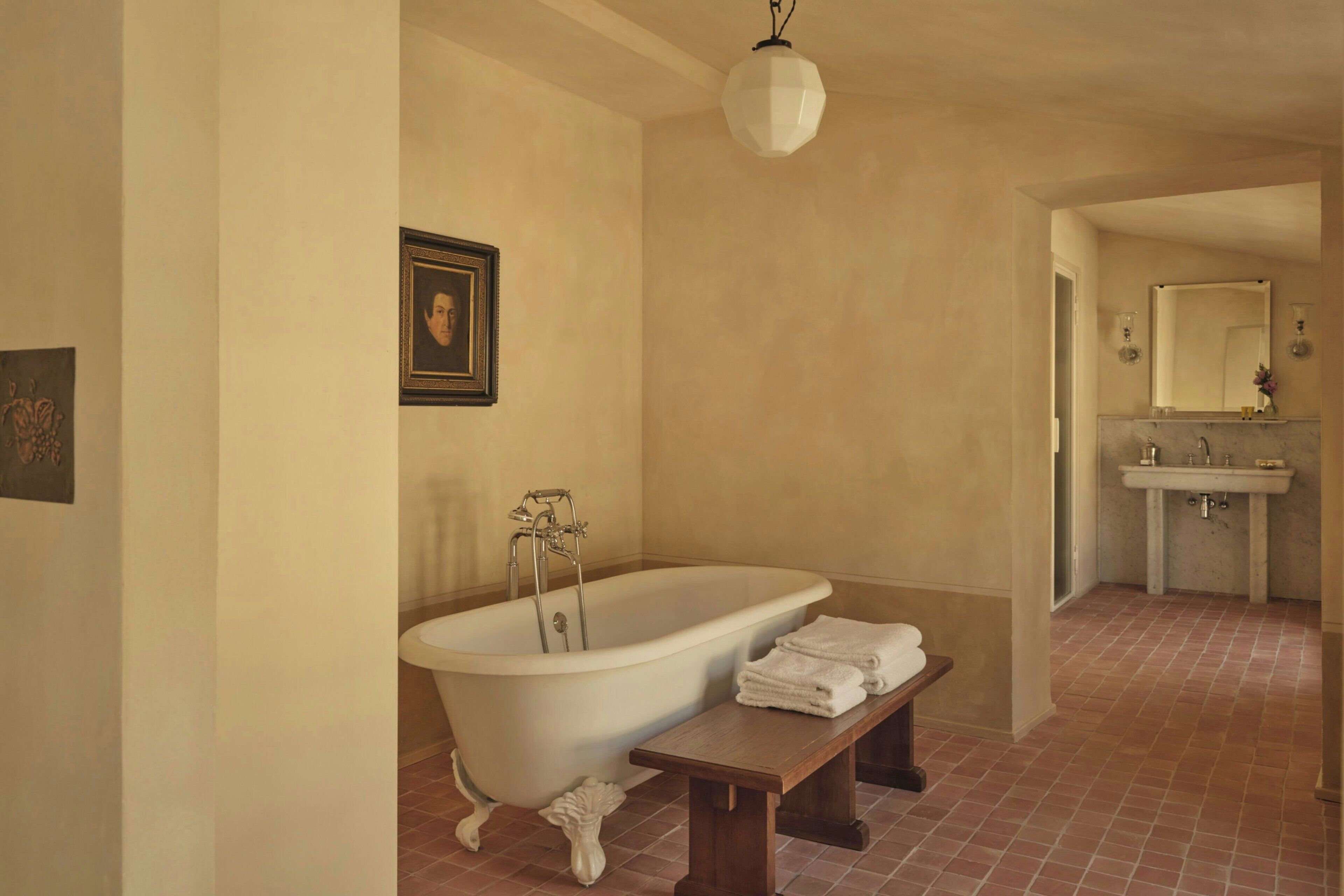 Une salle de bain avec une baignoire sur pieds, un banc, un tableau encadré au mur et un lavabo avec un miroir.