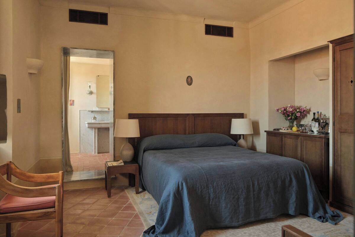 A bedroom with a bed, bedside tables, a chair, a desk, a wardrobe, and a bathroom visible through an open door.