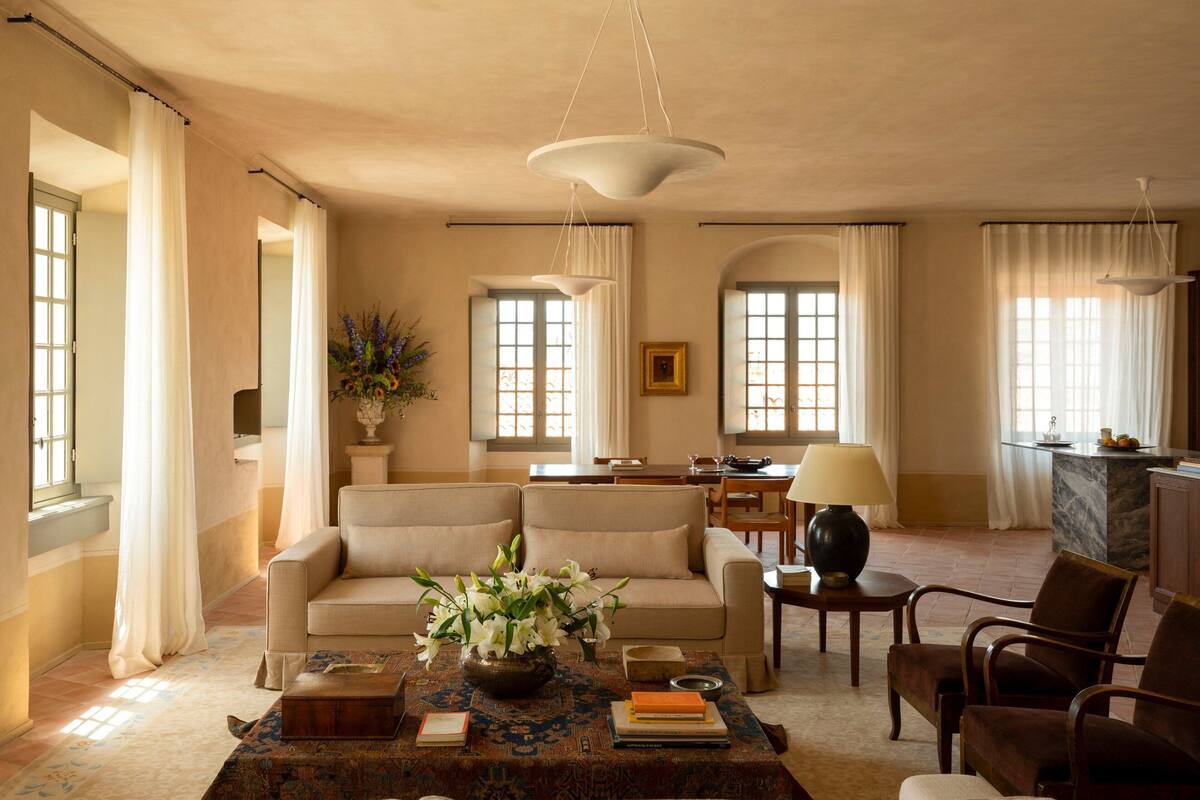 A spacious living room with a sofa, armchairs, a coffee table with books and a vase of flowers, and a dining area with a table and chairs.