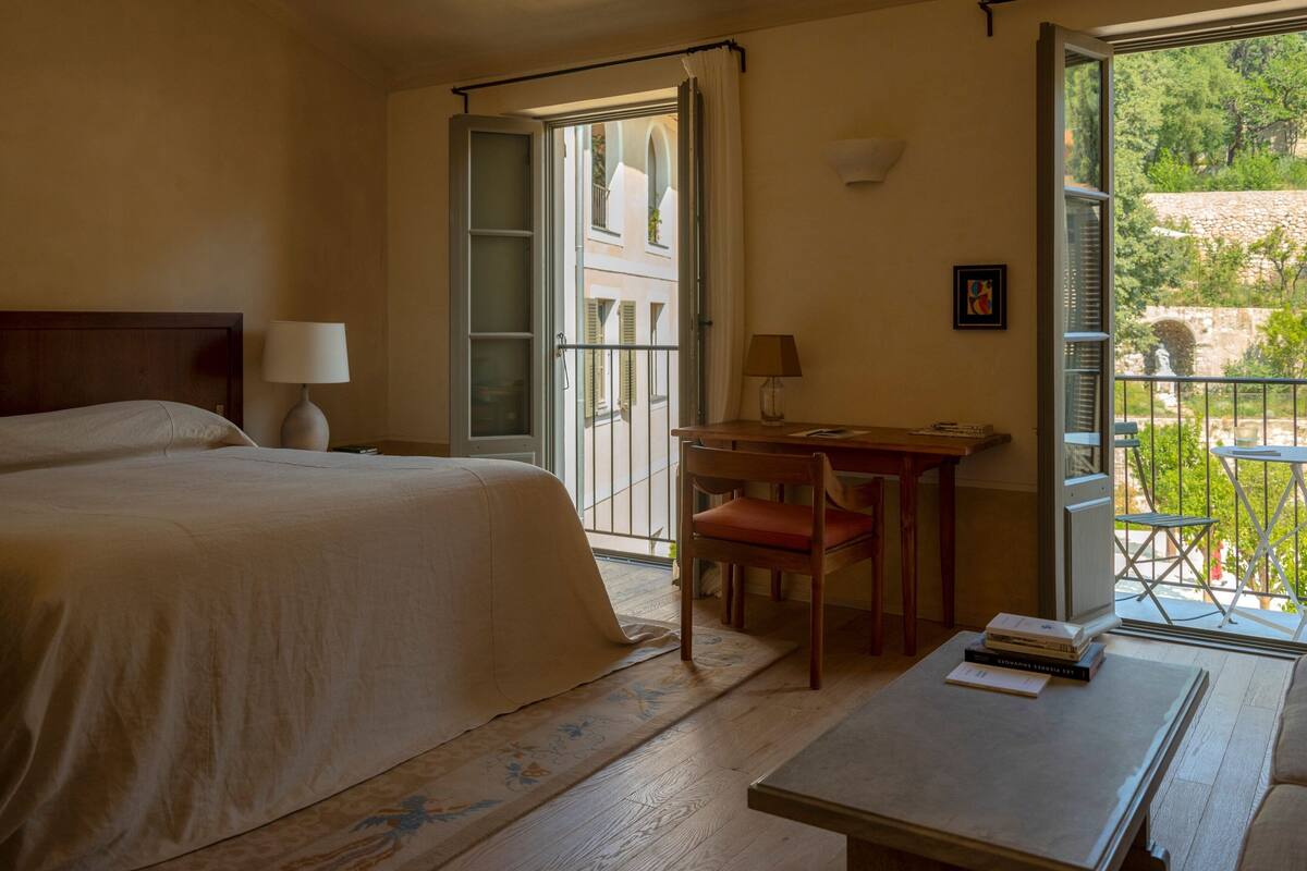 A hotel room with a bed, a desk, a chair, and a table. There is a window with a view of greenery outside.