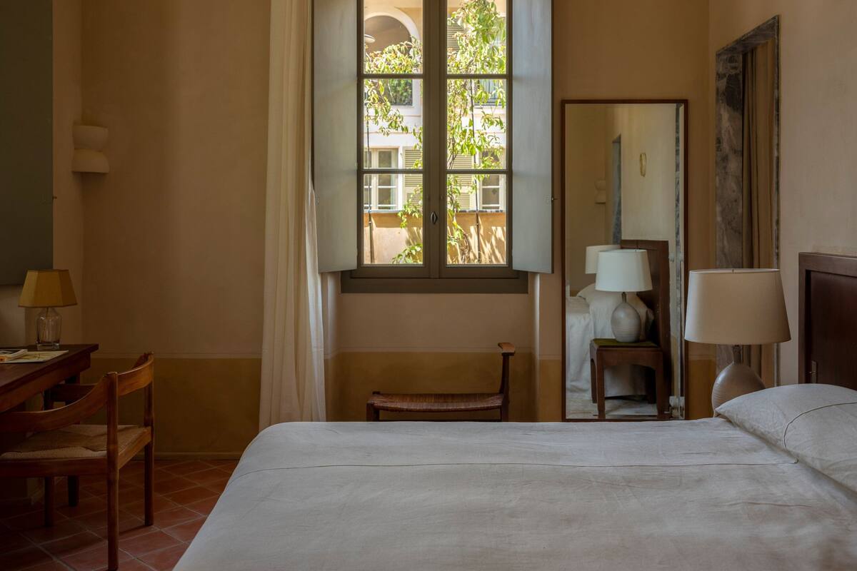 A hotel room with a bed, a desk, a chair, a window with a view of buildings, and a mirror.