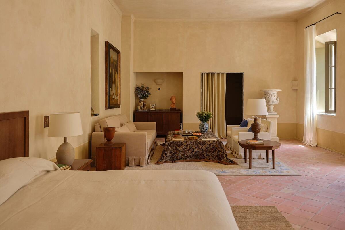 A hotel room with a large bed, a sofa, a small table, and a desk. There is a window with curtains, a painting on the wall, and a vase on the desk.