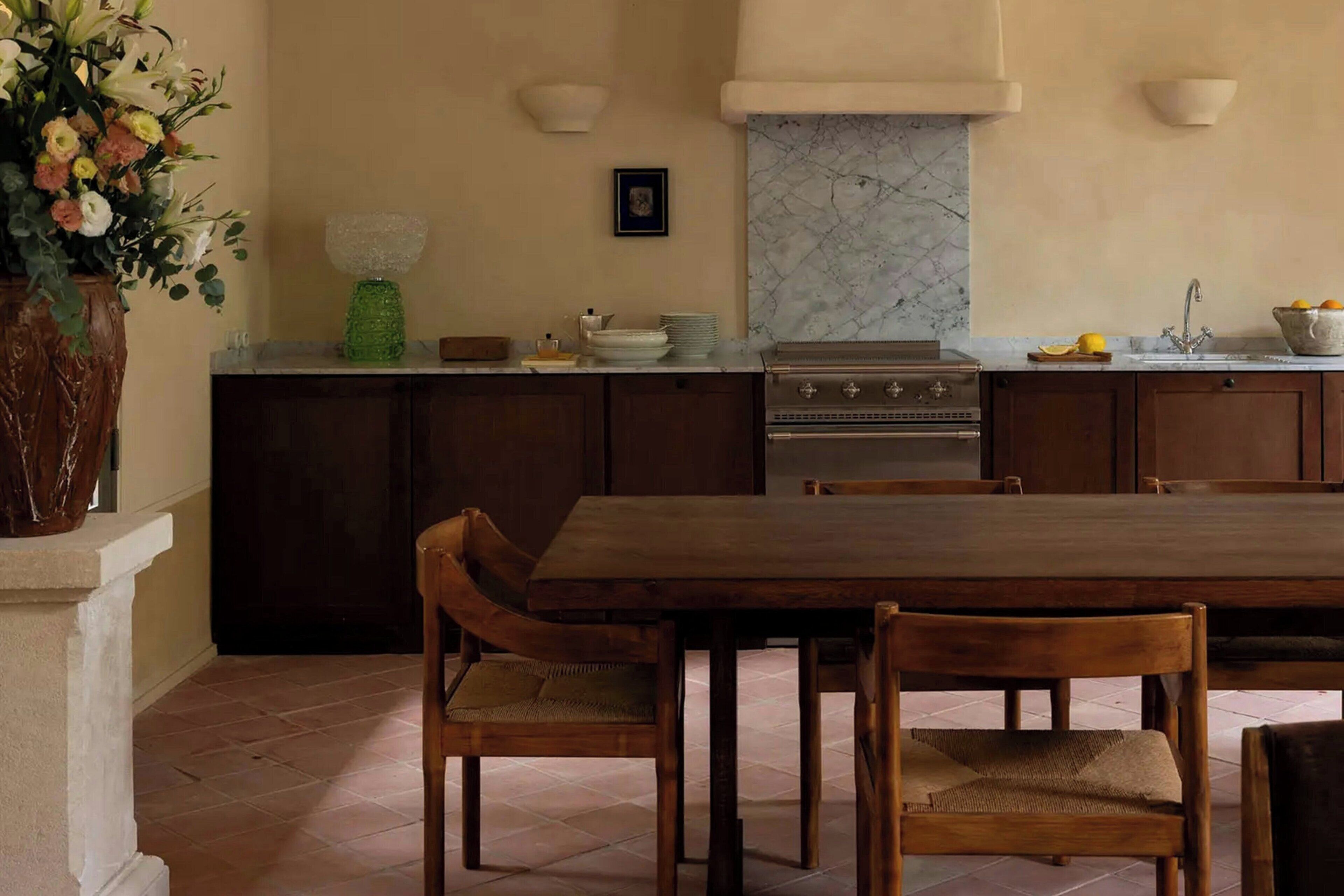 A kitchen with dark wooden cabinets, a marble backsplash, a wooden dining table with chairs, and a vase of flowers on the counter.