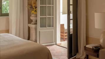 Une chambre d’hôtel avec un lit, une porte donnant sur un espace extérieur comprenant une chaise en osier et une table, ainsi qu’un vase avec des fleurs séchées.