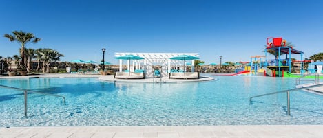 A clear blue pool with lounge chairs and umbrellas, a water play area, and a white pergola structure.