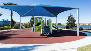 A playground with a blue canopy, red rubber flooring, and a water body in the background.