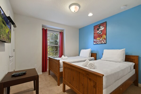 A hotel room with two wooden beds, a desk with a remote control, a window with red curtains, and a wall with a Mickey Mouse poster.