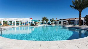 Une piscine d’un bleu éclatant, bordée de chaises longues et d’ombrelles, entourée de palmiers et d’une pergola blanche.