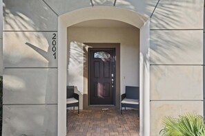L’entrée d’un immeuble avec une porte sombre, deux chaises en osier et un chemin pavé de briques.