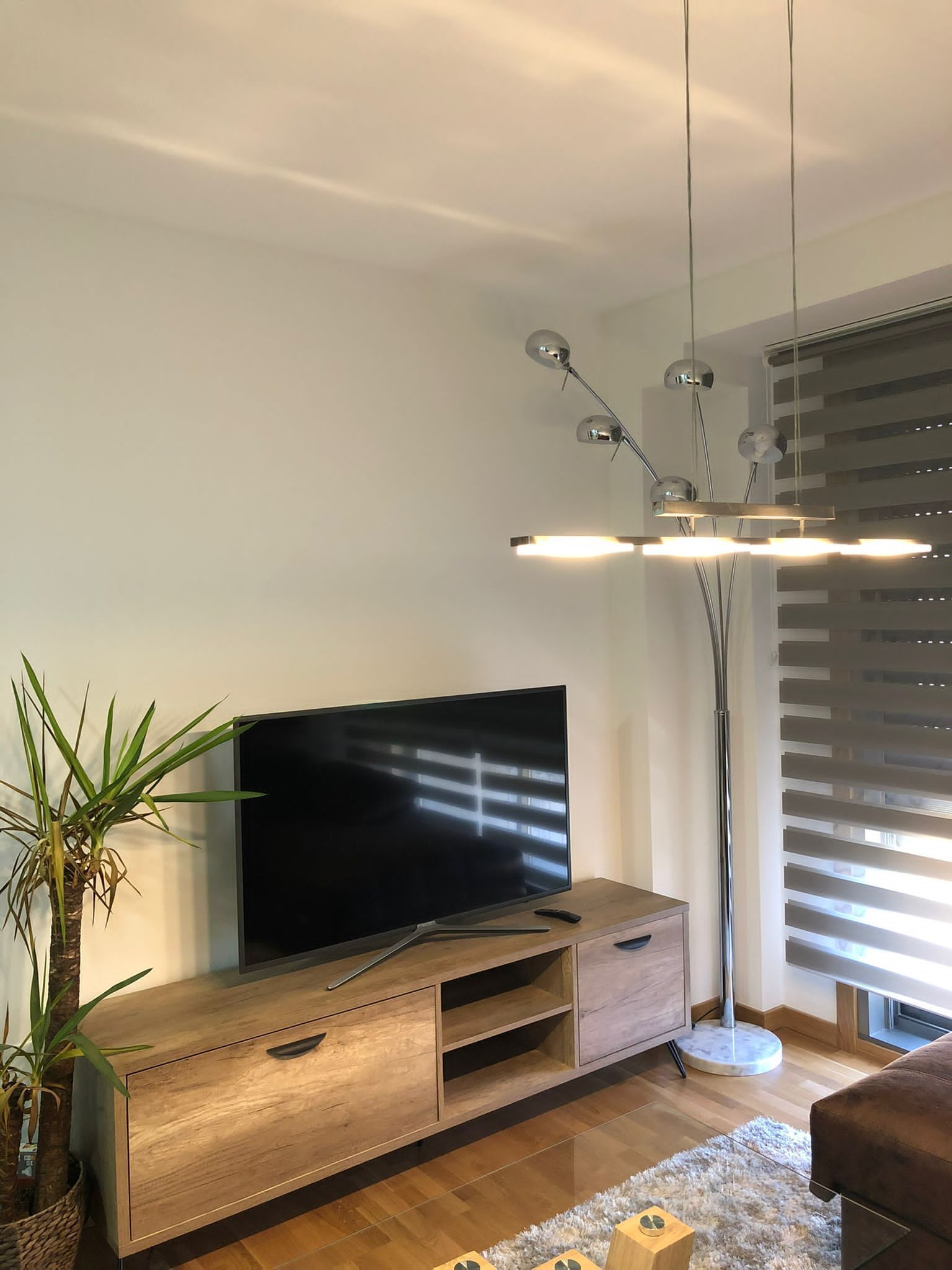 A modern living room with a flat-screen TV on a wooden TV stand, a tall floor lamp, and a large window with blinds.