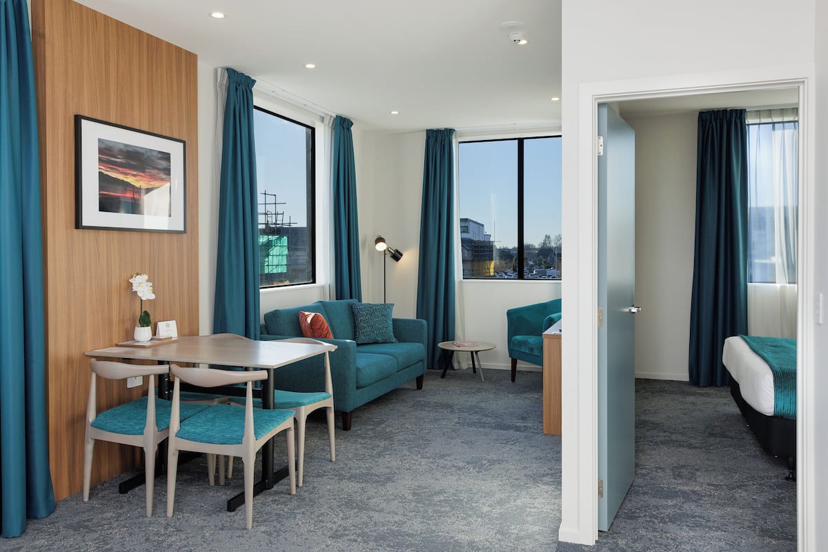 Une chambre d’hôtel moderne avec un mur d’accent en bois, des rideaux turquoise, un canapé, une table à manger avec des chaises, une petite table d’appoint et un lit recouvert d’une couverture turquoise.