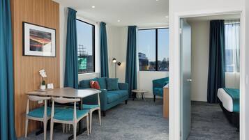 A modern hotel room with a wooden accent wall, teal curtains, a sofa, a dining table with chairs, a small side table, and a bed with a teal blanket.