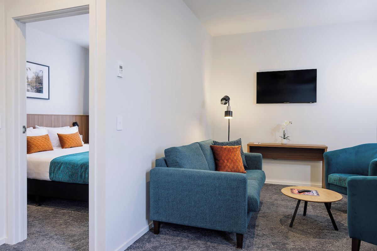 Una habitación de hotel moderna con una cama, dos sofás, una mesita, un televisor y una lámpara de pie.