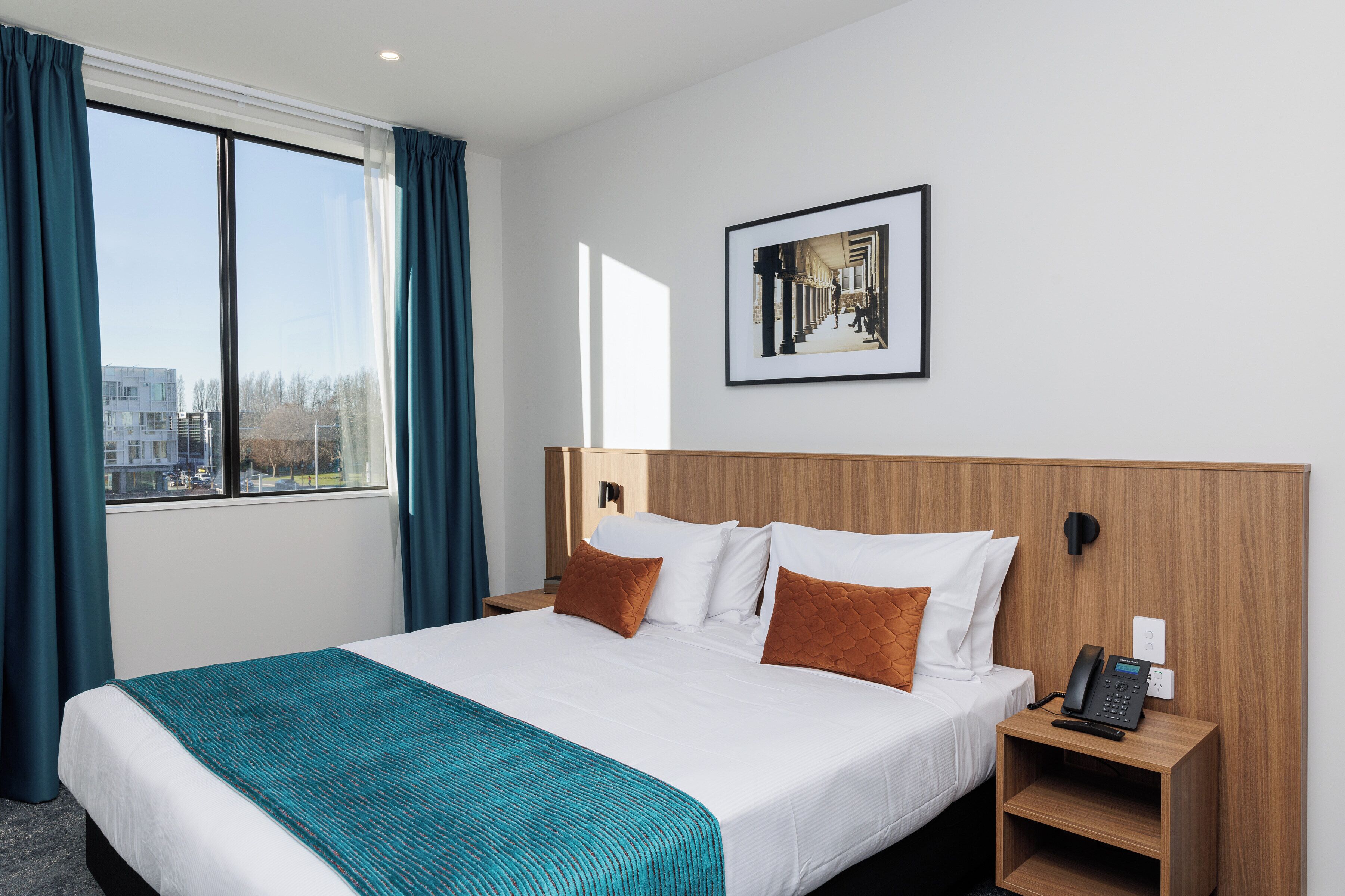 A hotel room with a large bed, a view of the city through the window, a framed picture on the wall, and a bedside phone.