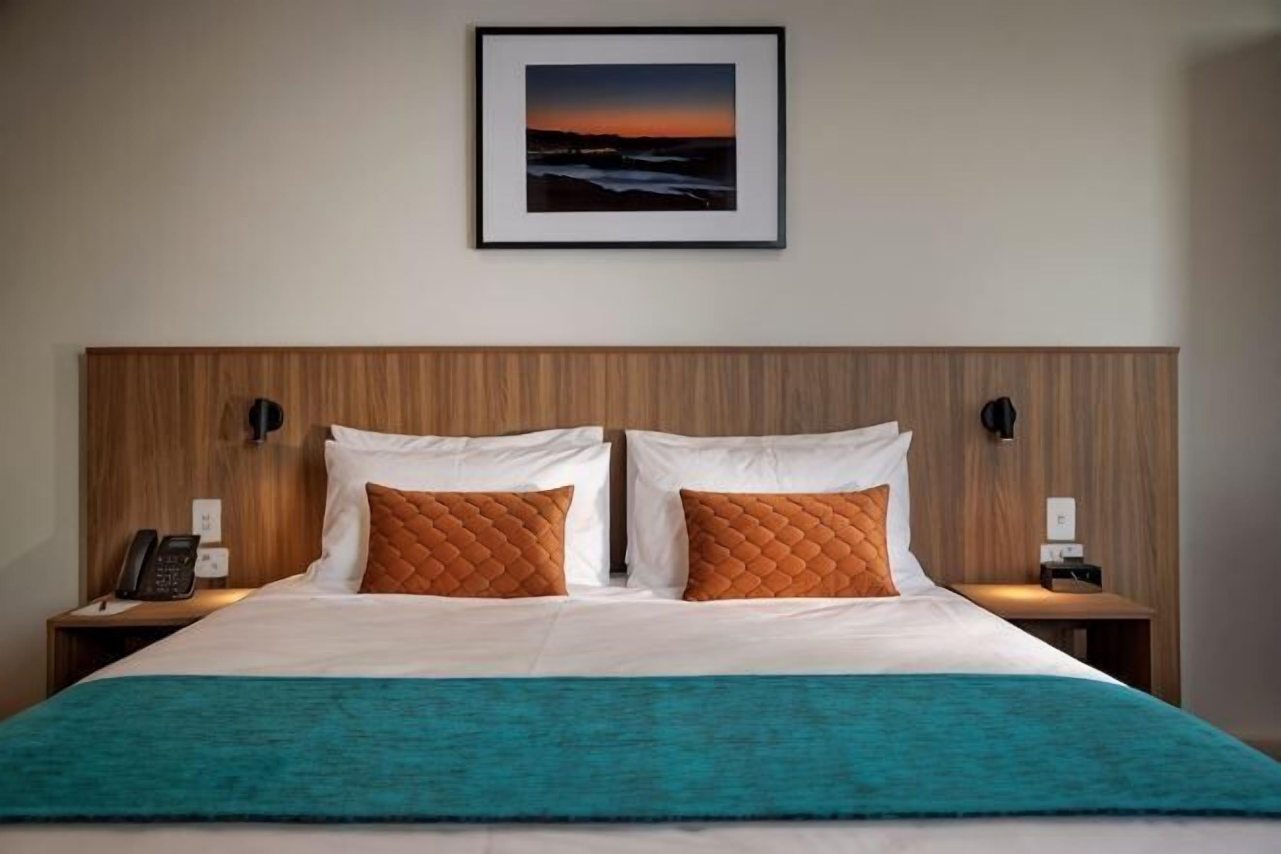 Ein Hotelzimmer mit einem Bett, zwei Nachttischen, einem Wandtelefon und einem gerahmten Bild über dem Bett.