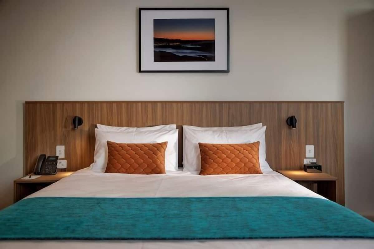 Une chambre d’hôtel comprenant un lit, deux tables de chevet, une petite table, un téléphone fixé au mur et un tableau encadré au-dessus du lit.