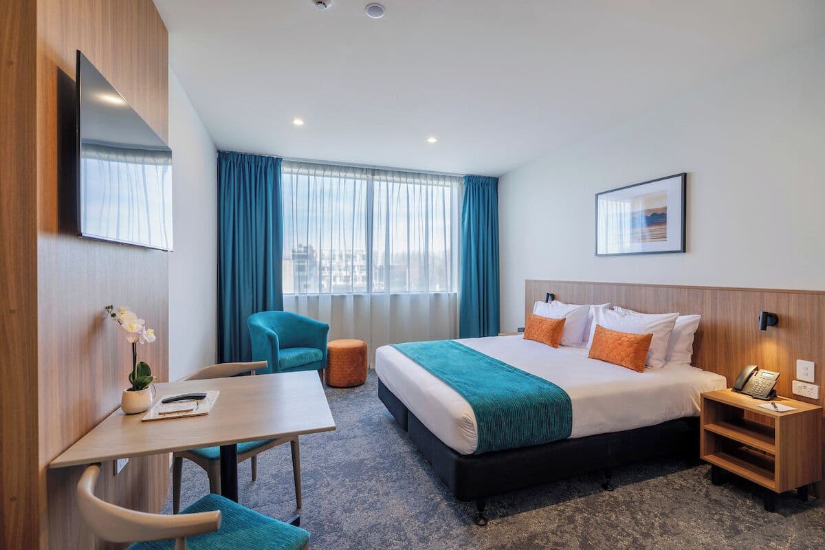 Une chambre d’hôtel comprenant un lit, un bureau, une chaise, une petite table, une table de chevet, un téléphone, un tableau encadré et une fenêtre avec des rideaux.
